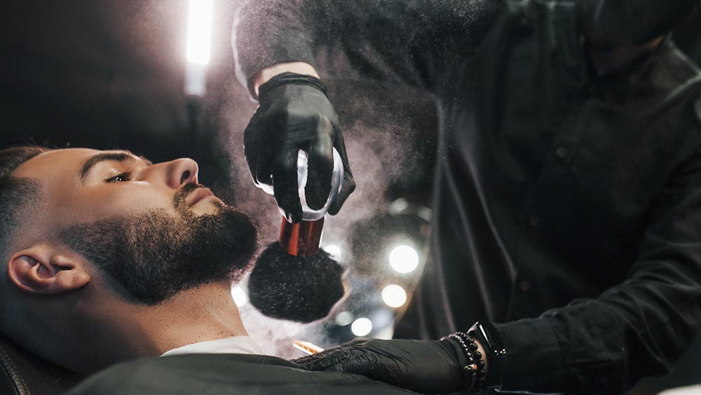 Joven con peinado elegante sentado y afeitándose la barba en la peluquería